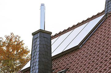 Solarpanele auf dem mit roten Pfannen eingedeckten Dach eines Wohnhauses. Im Vordrgrund ein Schornstein.